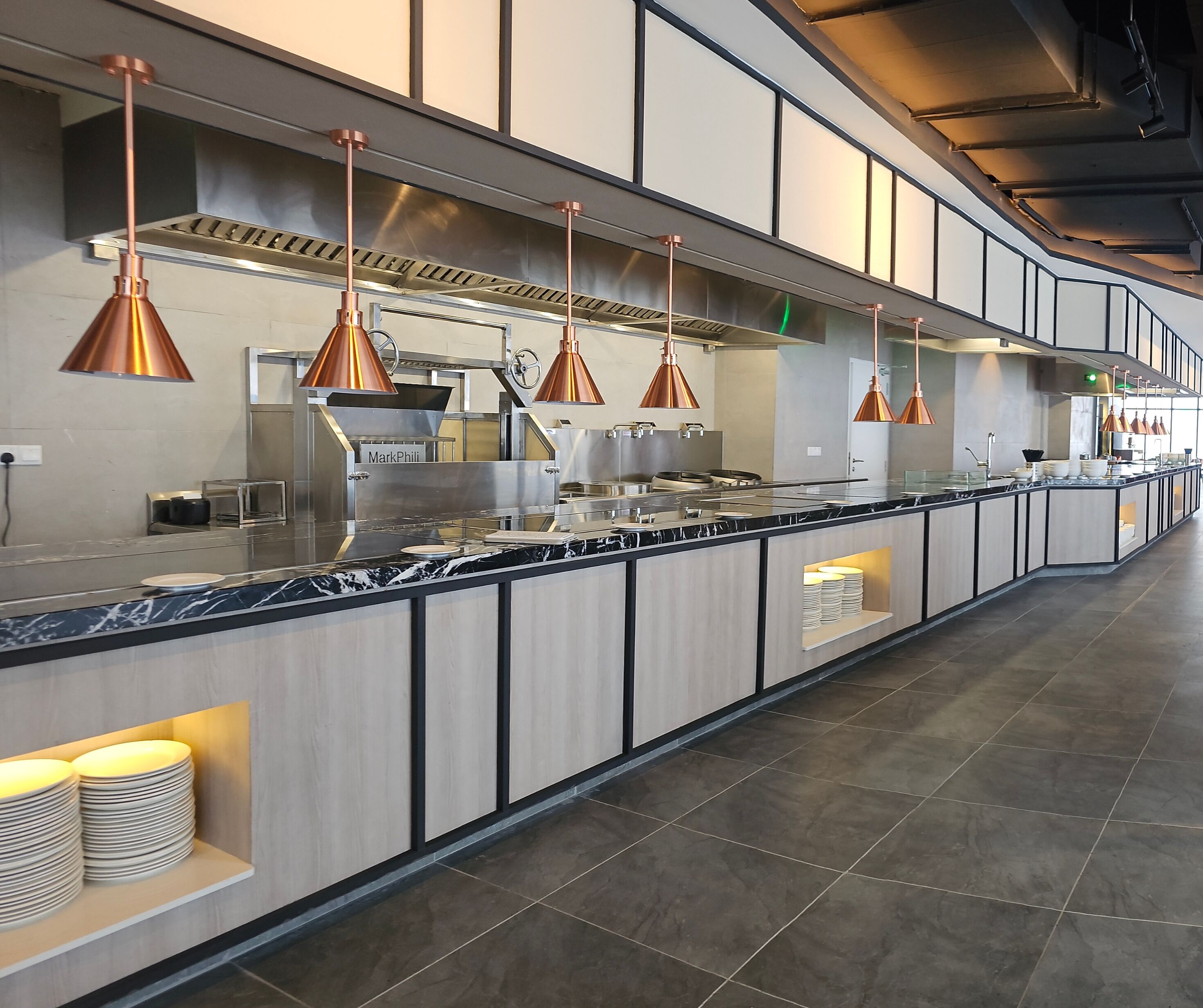 A modern kitchen with a long counter, copper pendant lights, and stainless steel appliances.