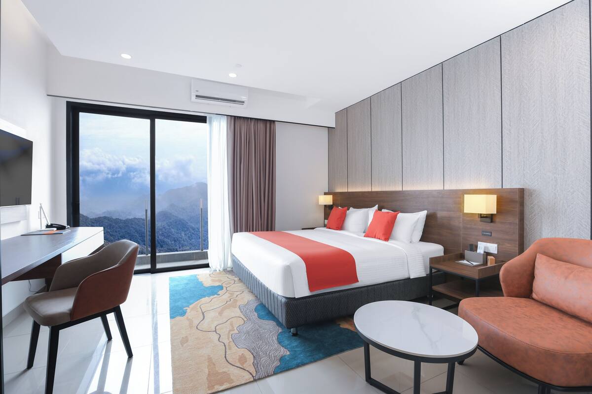 Habitación de hotel moderna con una cama grande, un escritorio, una silla, una mesita y vistas a las montañas a través de un ventanal amplio.