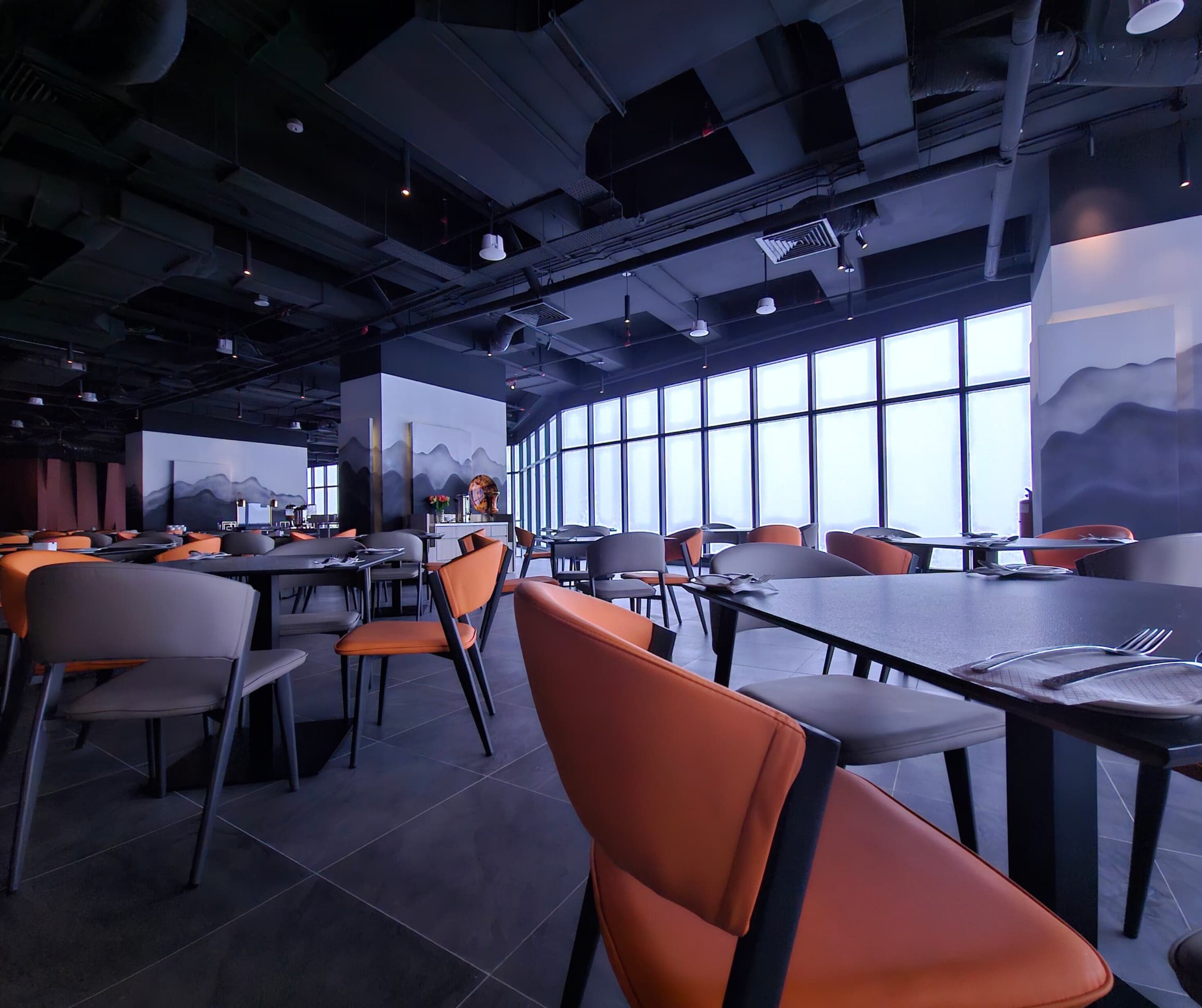Een modern restaurantinterieur met hoge plafonds, open ventilatiekanalen en grote ramen met uitzicht op het omliggende landschap. Tafels en stoelen staan klaar voor de gasten.
