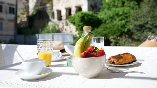 Desayuno con una taza, un vaso de jugo de naranja, un tazón de fresas y plátanos, y un croissant sobre una mesa blanca.