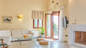 Ein geräumiges Wohnzimmer mit weißem Sofa, einem Glastisch, einem Kamin und einem großen Fenster mit Vorhängen.