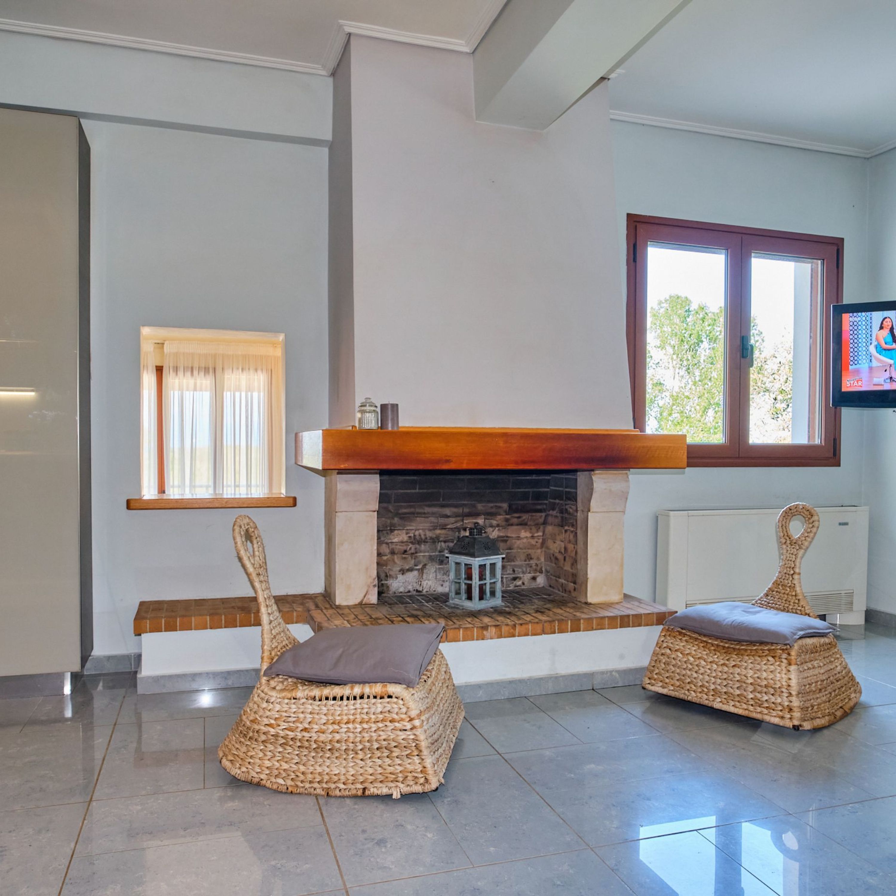 A room with a fireplace, two wicker chairs with cushions, a window with curtains, and a television mounted on the wall.