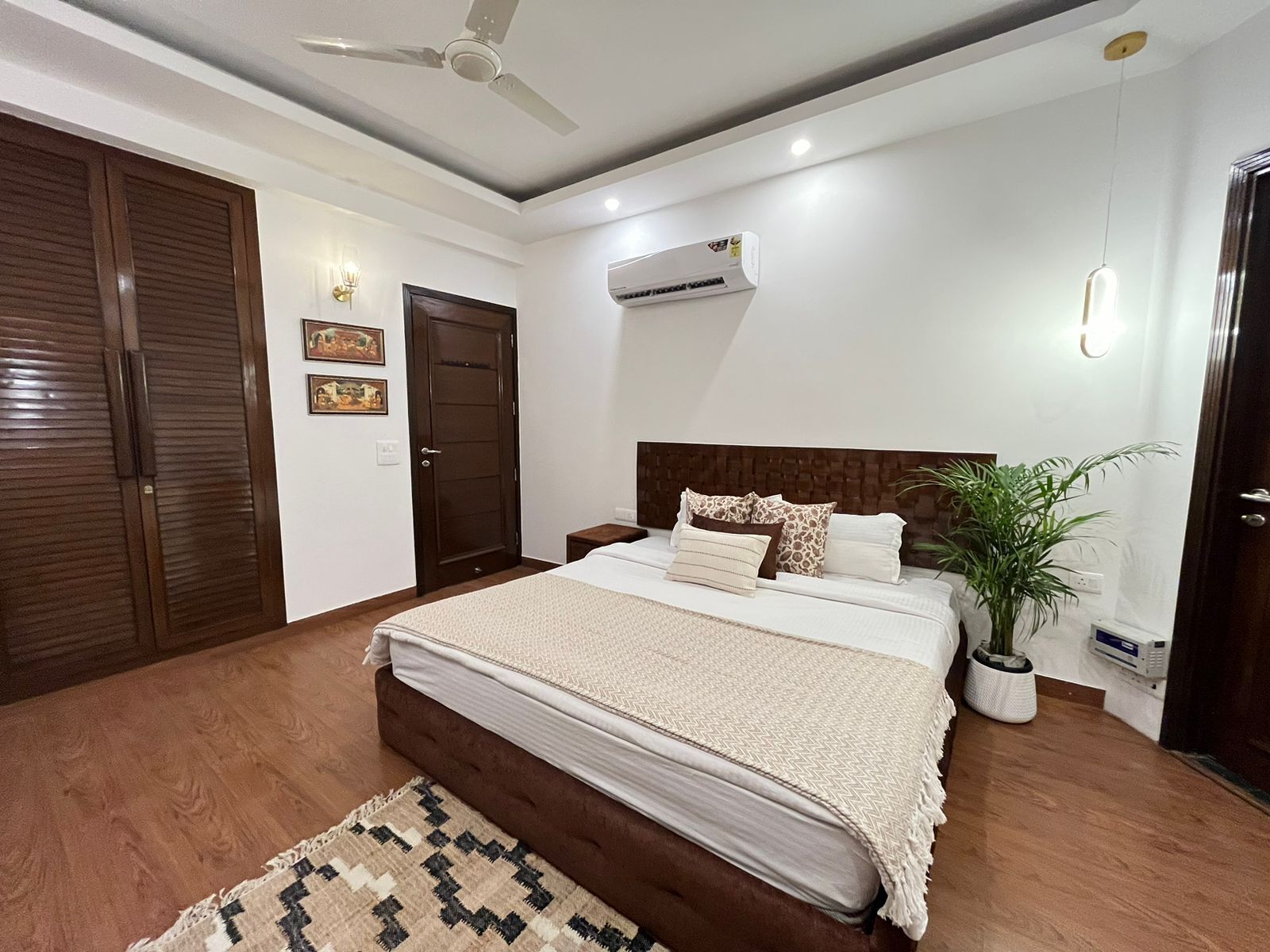 A bedroom with a bed, a ceiling fan, an air conditioning unit, a potted plant, and a wooden floor.