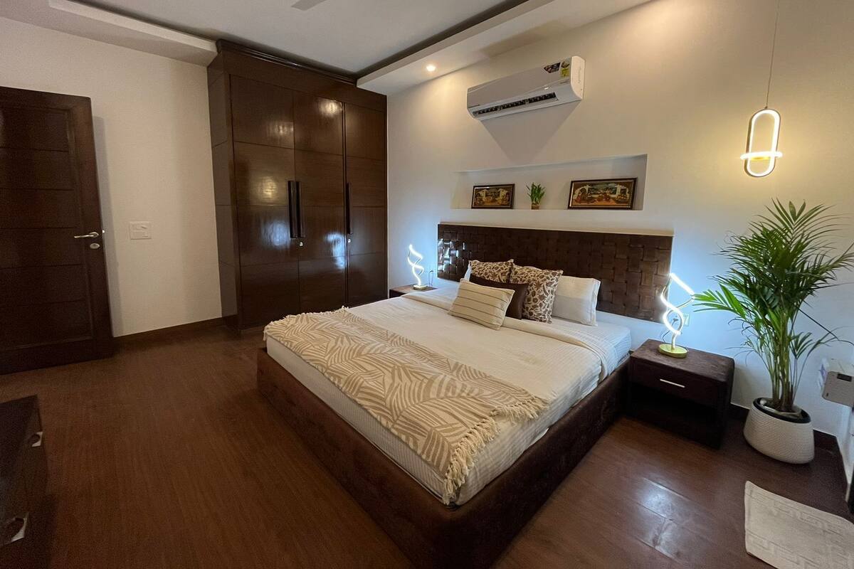 Ein Schlafzimmer mit einem großen Bett, einem Kleiderschrank, einem Deckenventilator, einer Klimaanlage, einem Nachttisch mit Lampe und einer Topfpflanze.