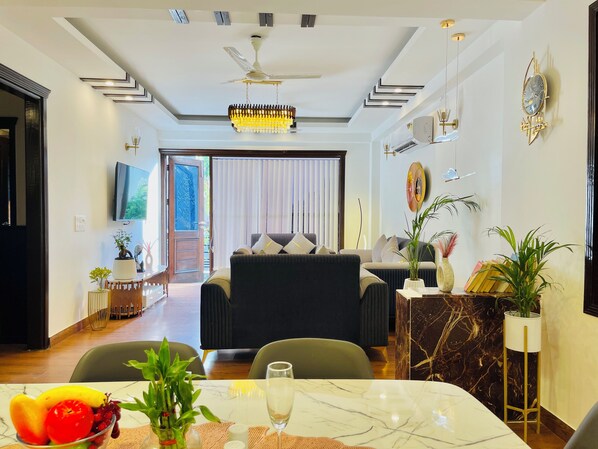 A modern living room with a sofa, a dining table with fruit and a glass, and a TV mounted on the wall.