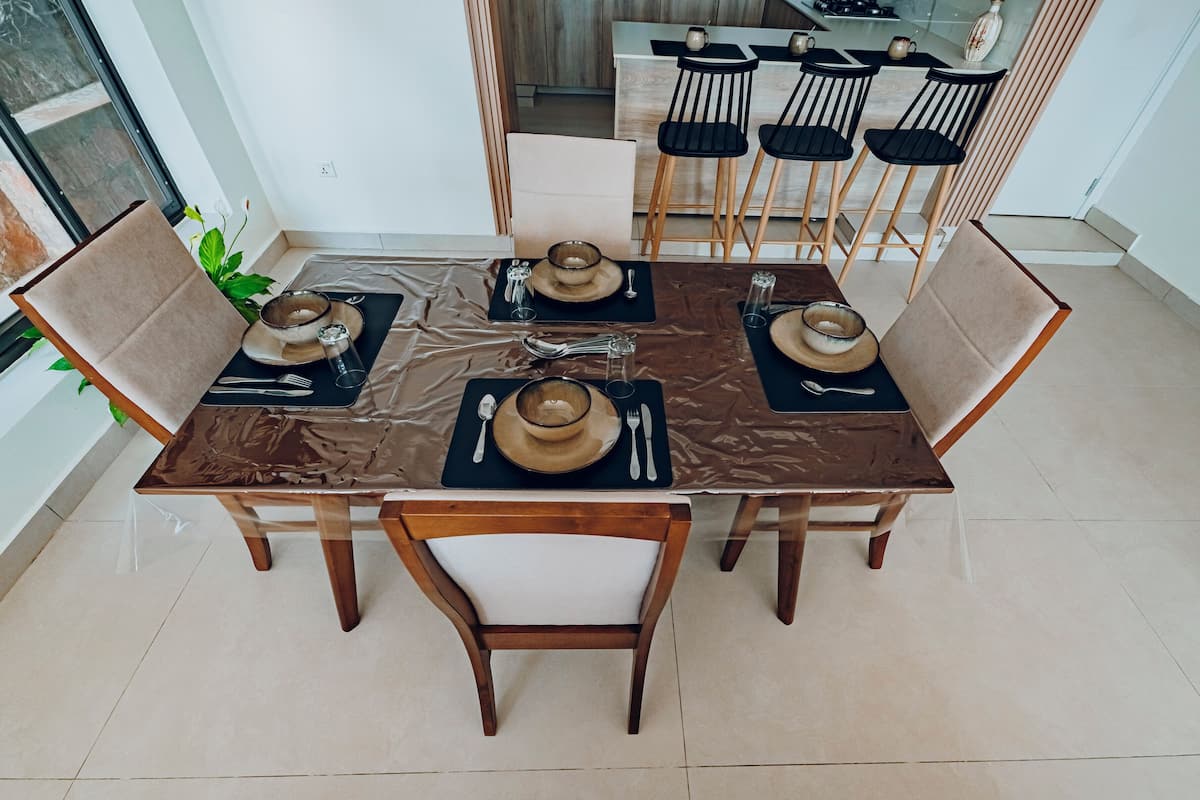 A dining table set for four with wooden chairs, a kitchen in the background, and a plant on the left.