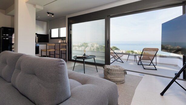 A modern living room with a sofa, a dining area with chairs, a small table, and a balcony with a view of the sea. - Athens Riviera Suites (Saronikos)