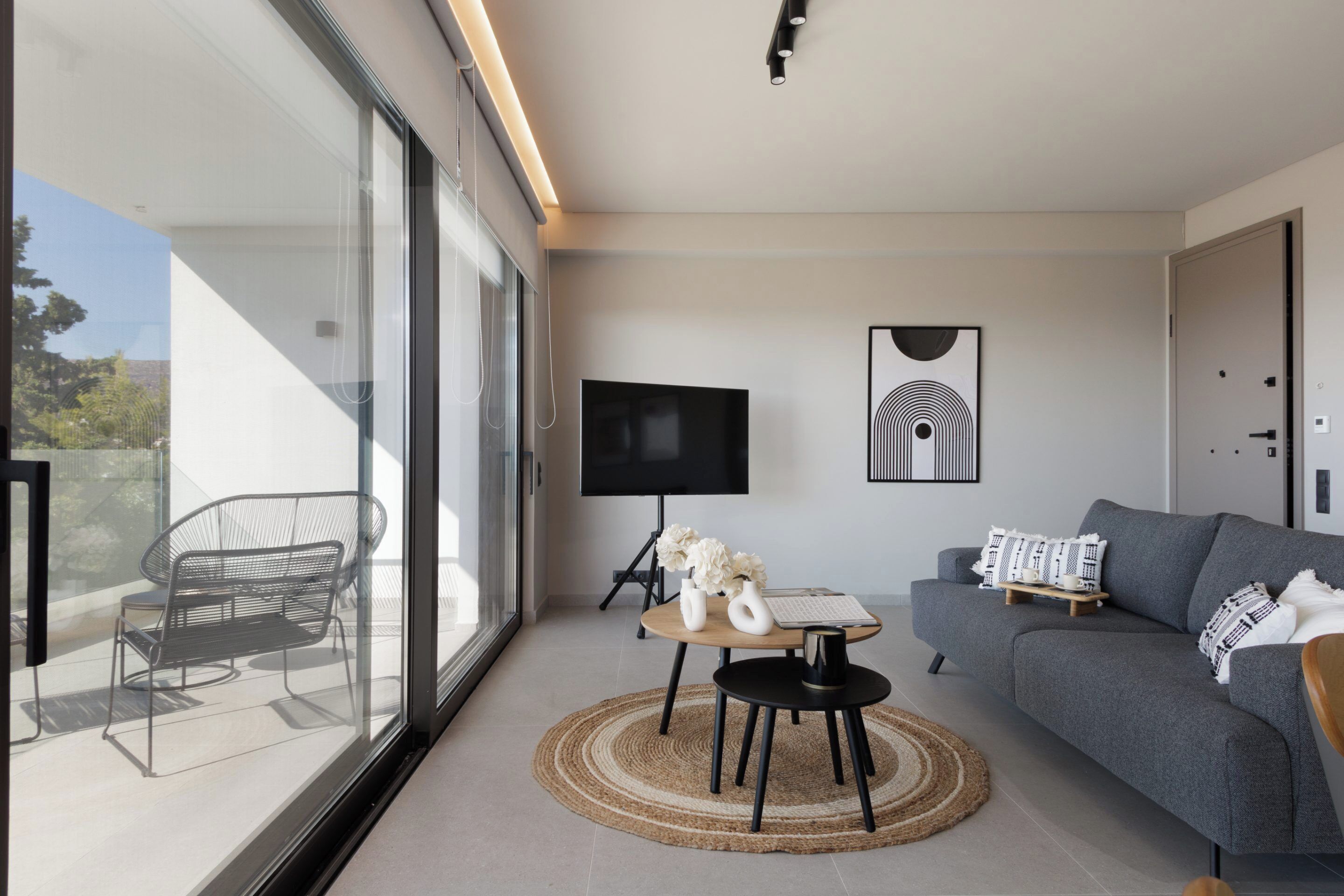 Un salon moderne avec un canapé gris, un téléviseur posé sur un meuble, une table ronde agrémentée d’un vase et d’un clavier, et une grande fenêtre donnant sur une chaise placée à l’extérieur.