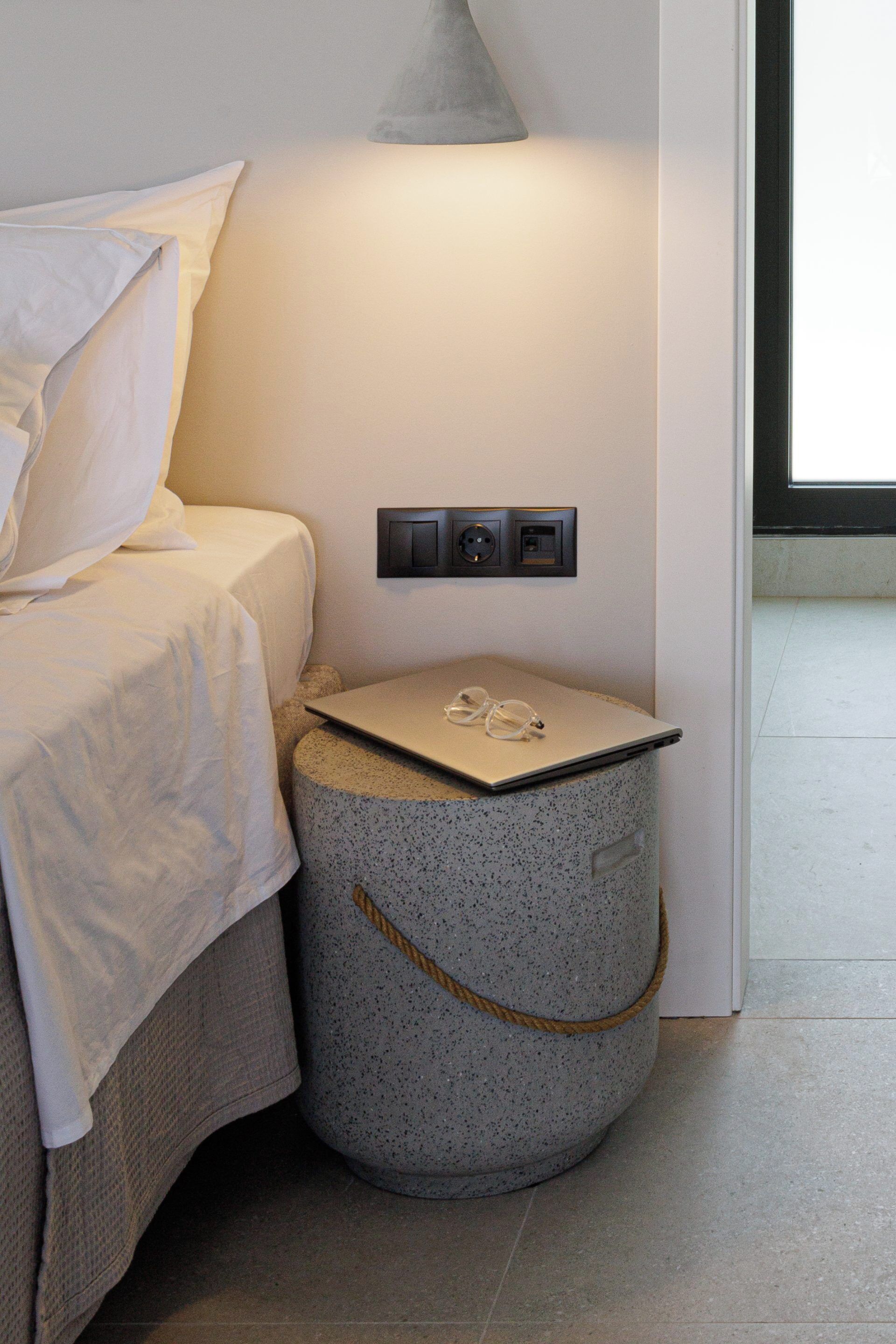 Un dormitorio con cama, una mesita redonda con una computadora portátil y vasos, una lámpara y un enchufe.