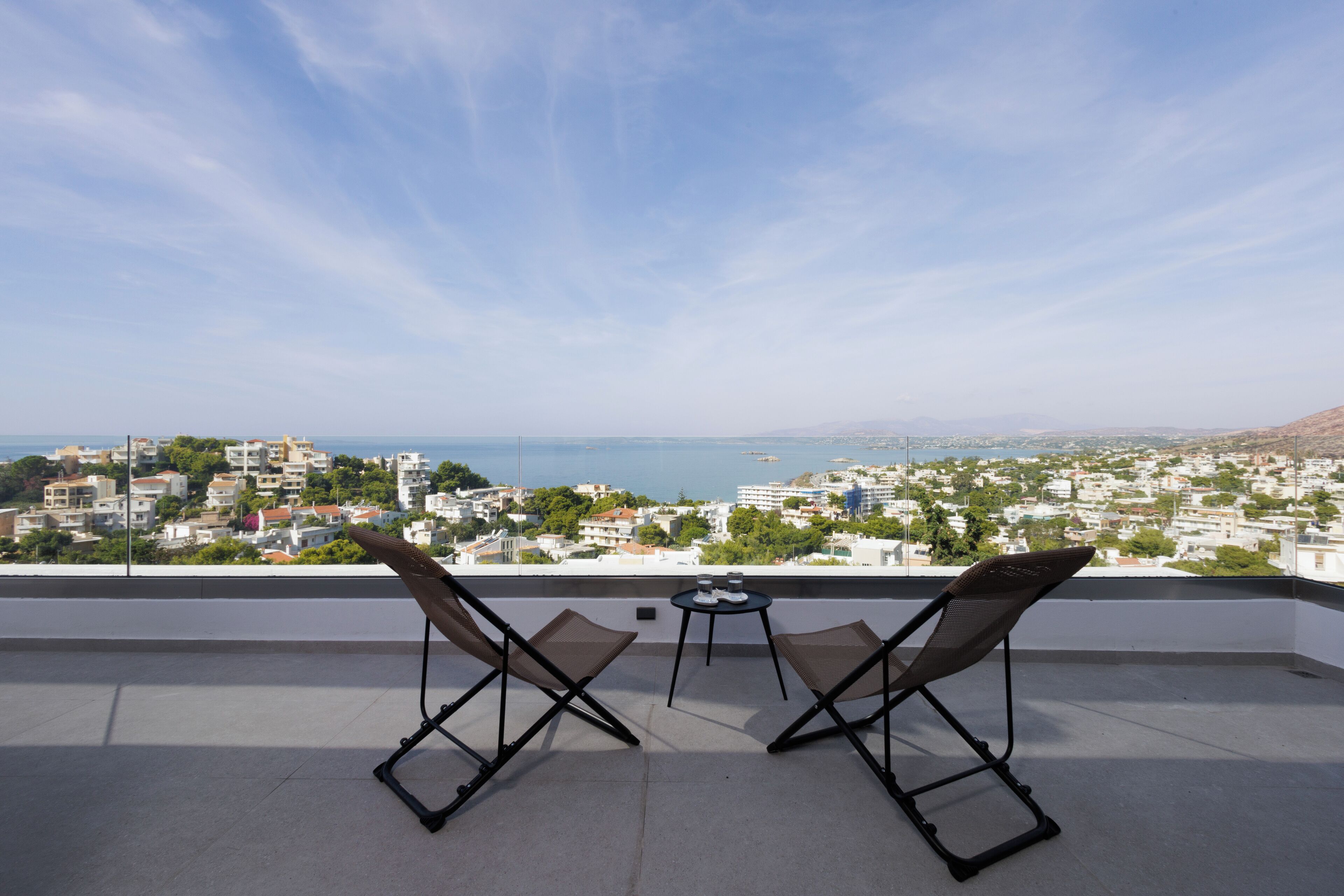 A balcony with two chairs and a small table, overlooking a cityscape with buildings and a body of water.