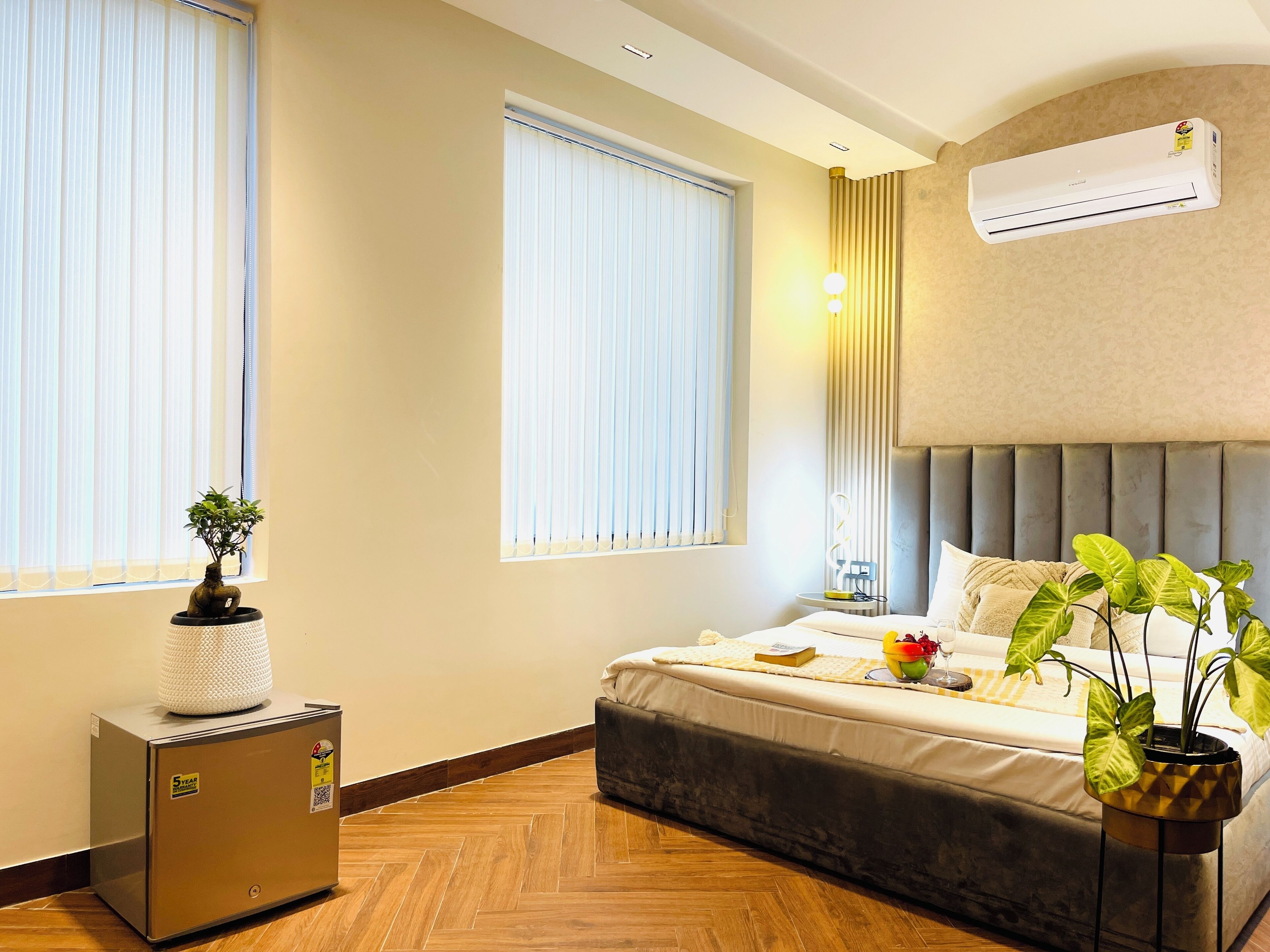 A hotel room with a bed, a bedside table, a potted plant, a heater, and a window with blinds.