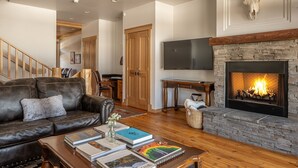 A living room with a leather sofa, a coffee table with books, a fireplace, and a mounted TV. - Cross-Country Ski Out the Back Door (Big Sky)
