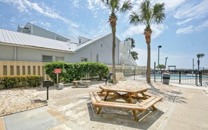 Un espace aménagé au bord de la piscine, avec une table et des bancs en bois, des palmiers et un bâtiment à la façade blanche.