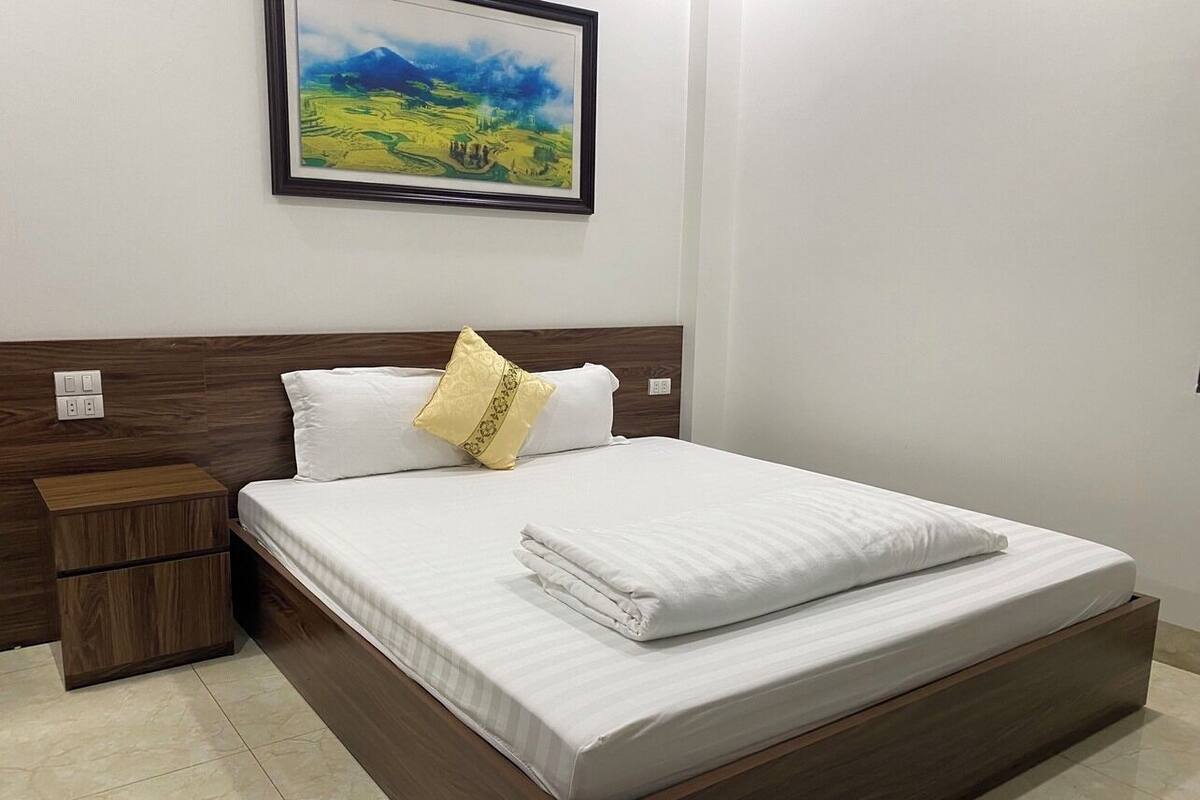 A neatly made bed with white linens and a single decorative pillow, a wooden nightstand with a drawer, and a framed landscape painting on the wall.