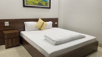 A neatly made bed with white linens and a single decorative pillow, a wooden nightstand with a drawer, and a framed landscape painting on the wall.