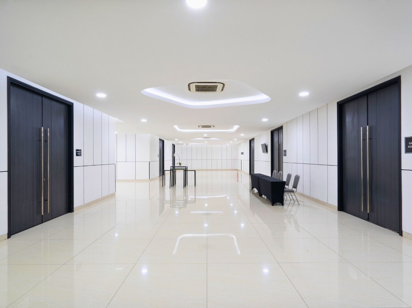 Un pasillo moderno de hotel con piso de baldosas brillantes, paredes en tonos oscuros y claros, y un área de recepción con mesa y sillas.