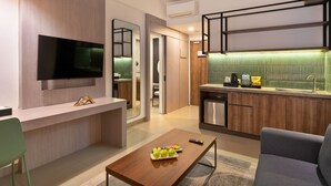 A modern living room with a flat-screen TV, a wooden coffee table with fruit, a sofa, and a kitchen area with a green backsplash and stainless steel appliances. - Laska Hotel Subang (Subang)