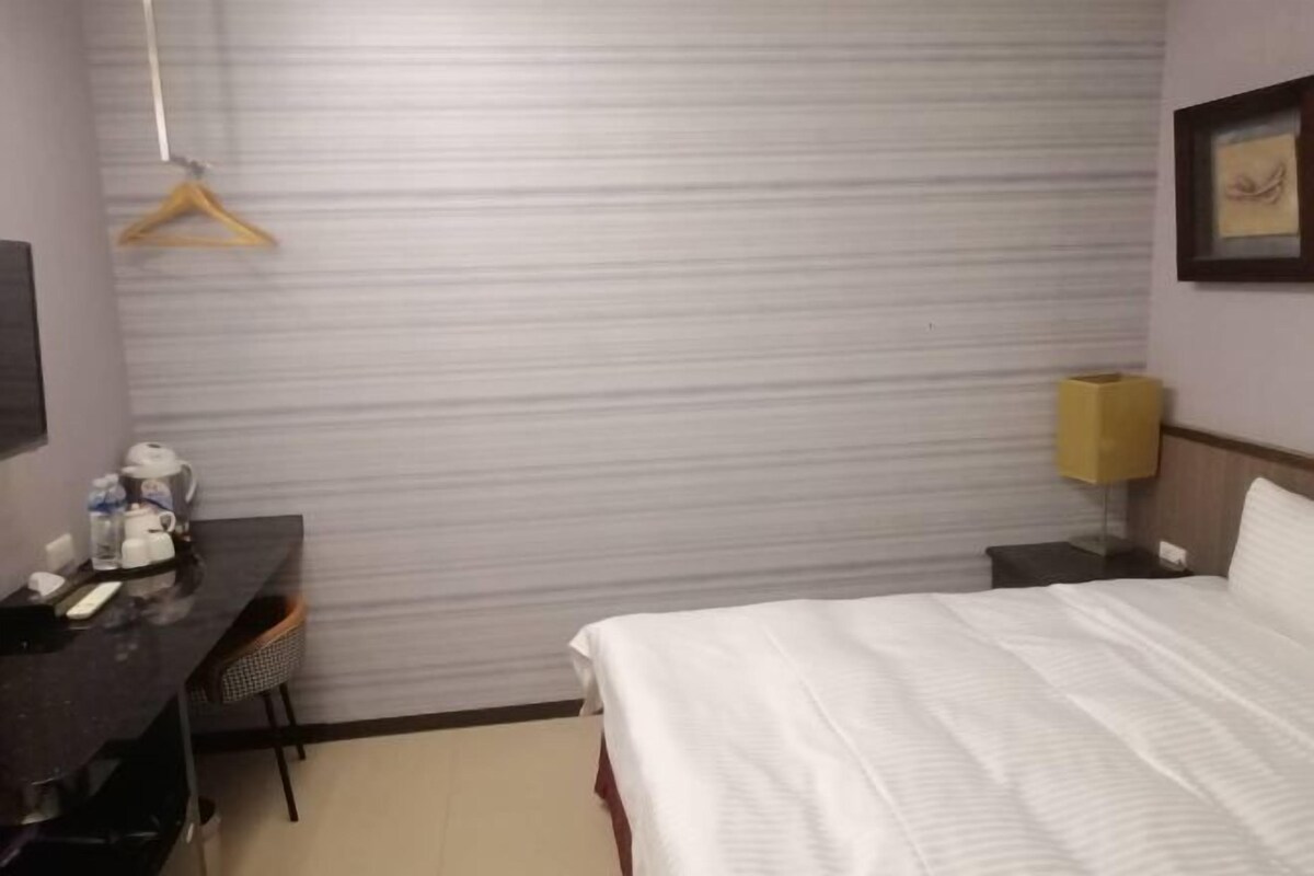Hotellihuone, jossa on sänky, työpöytä tuolilla, seinälle ripustettu naulakko, yöpöytä lampun kanssa ja seinällä oleva kehystetty kuva.