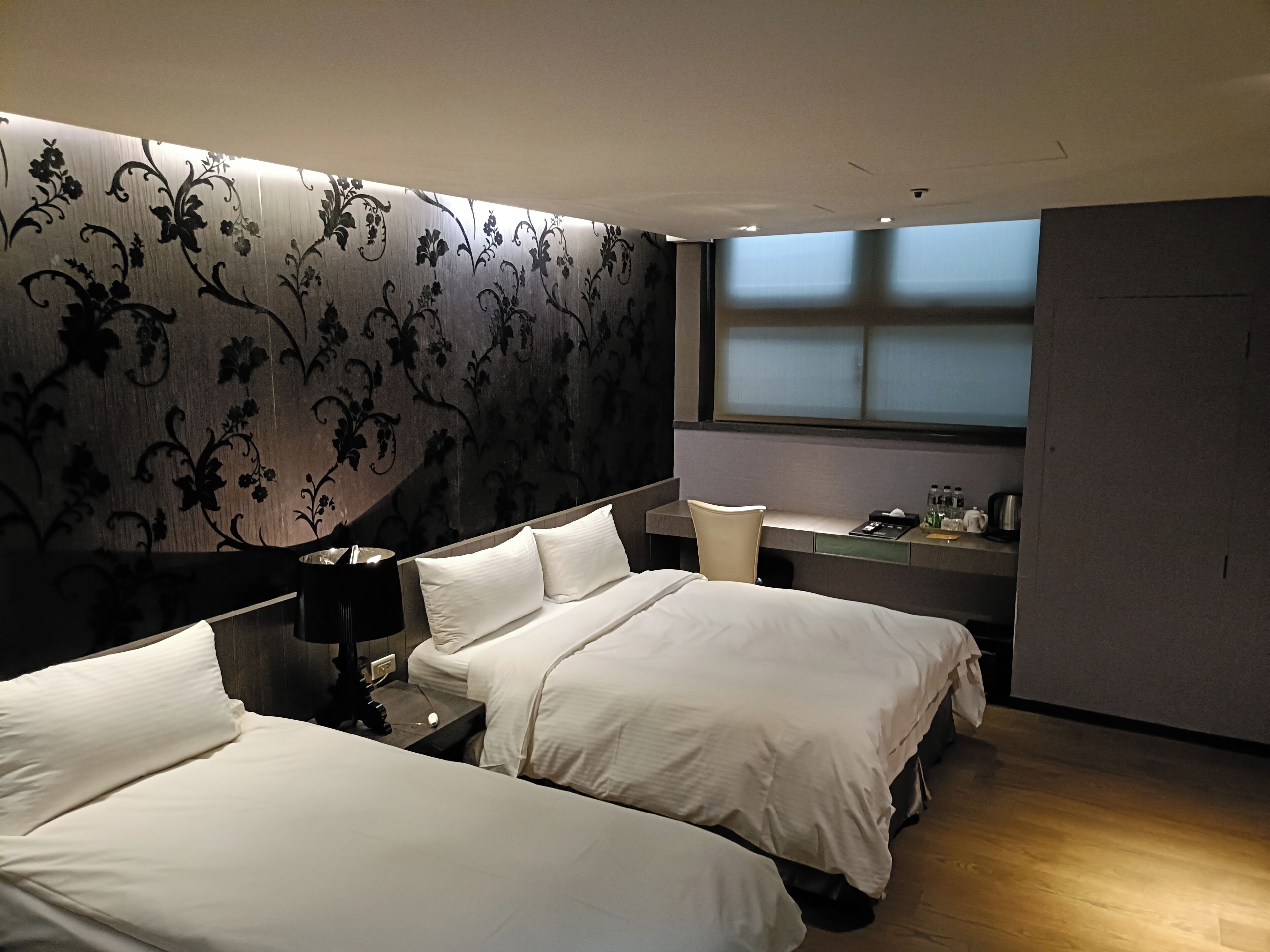 Habitación de hotel con dos camas, un escritorio, una silla y una ventana con persianas.