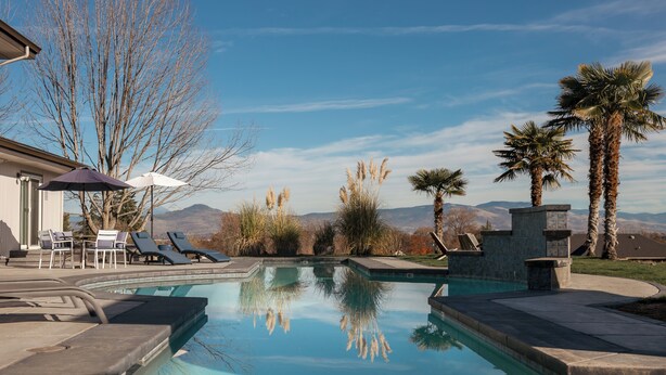Piscina con sillones de descanso y sombrillas, rodeada de palmeras y vistas a montañas.