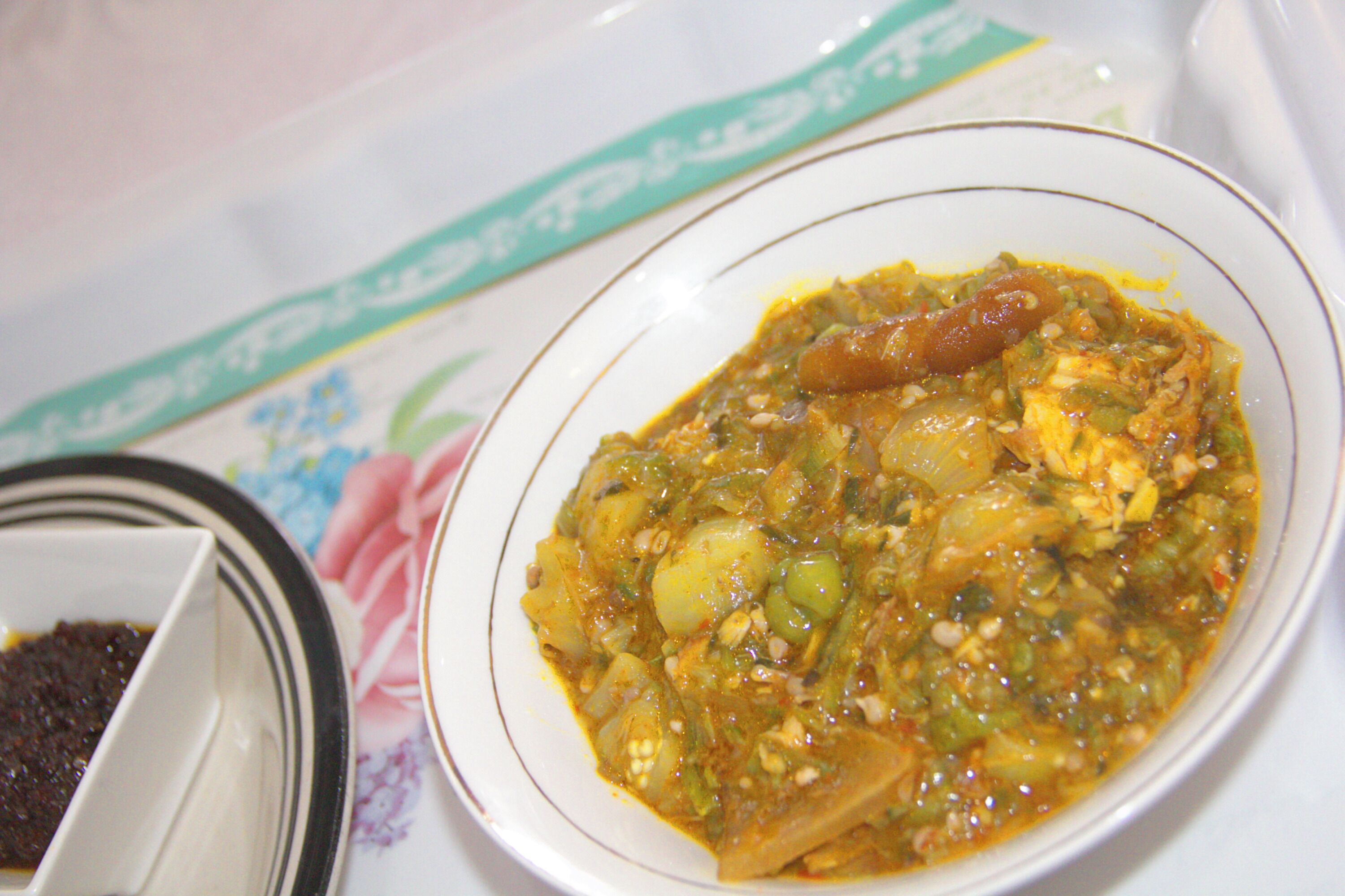A bowl of stew with visible ingredients like tomatoes and green peppers, served on a white plate with a floral tablecloth underneath.