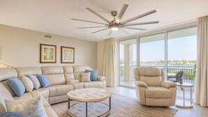 A spacious living room with a beige leather sofa, a round coffee table, and a beige armchair. There is a ceiling fan, large windows with curtains, and a view of the outdoors.