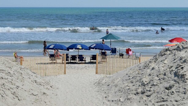 A beach with umbrellas and beach chairs, a wooden fence, and the ocean in the background.