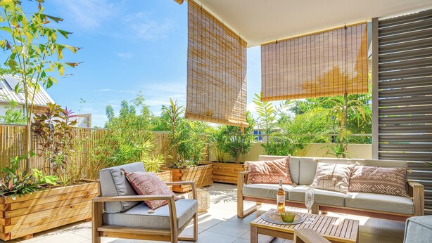Un patio con mobiliario de madera, incluyendo dos sofás y una silla, rodeado de plantas en macetas y una valla de bambú.
