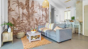 Un salón con un sofá gris, una pequeña mesa de centro con fruta, una lámpara trenzada y un mural tropical en la pared.