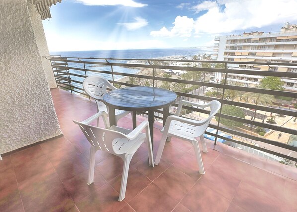 A balcony with a round table and white plastic chairs, overlooking a coastal area with buildings and a clear sky.