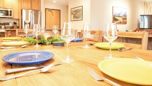 Une table à manger dressée avec des assiettes, des couverts et des verres à vin, entourée de chaises en bois dans une pièce bien éclairée, où l’on aperçoit des appareils de cuisine et des œuvres d’art accrochées aux murs.