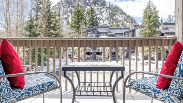 Un balcon avec deux chaises à motifs et une table, deux verres à vin, et une vue sur une montagne et des bâtiments.