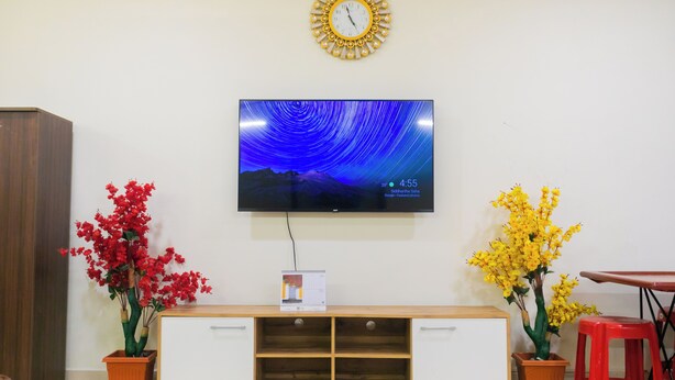 A room with a TV mounted on the wall, a wooden cabinet, two potted plants, and a red stool. - Roscotel Bay Stays 2BHK Near White Town (Puducherry)