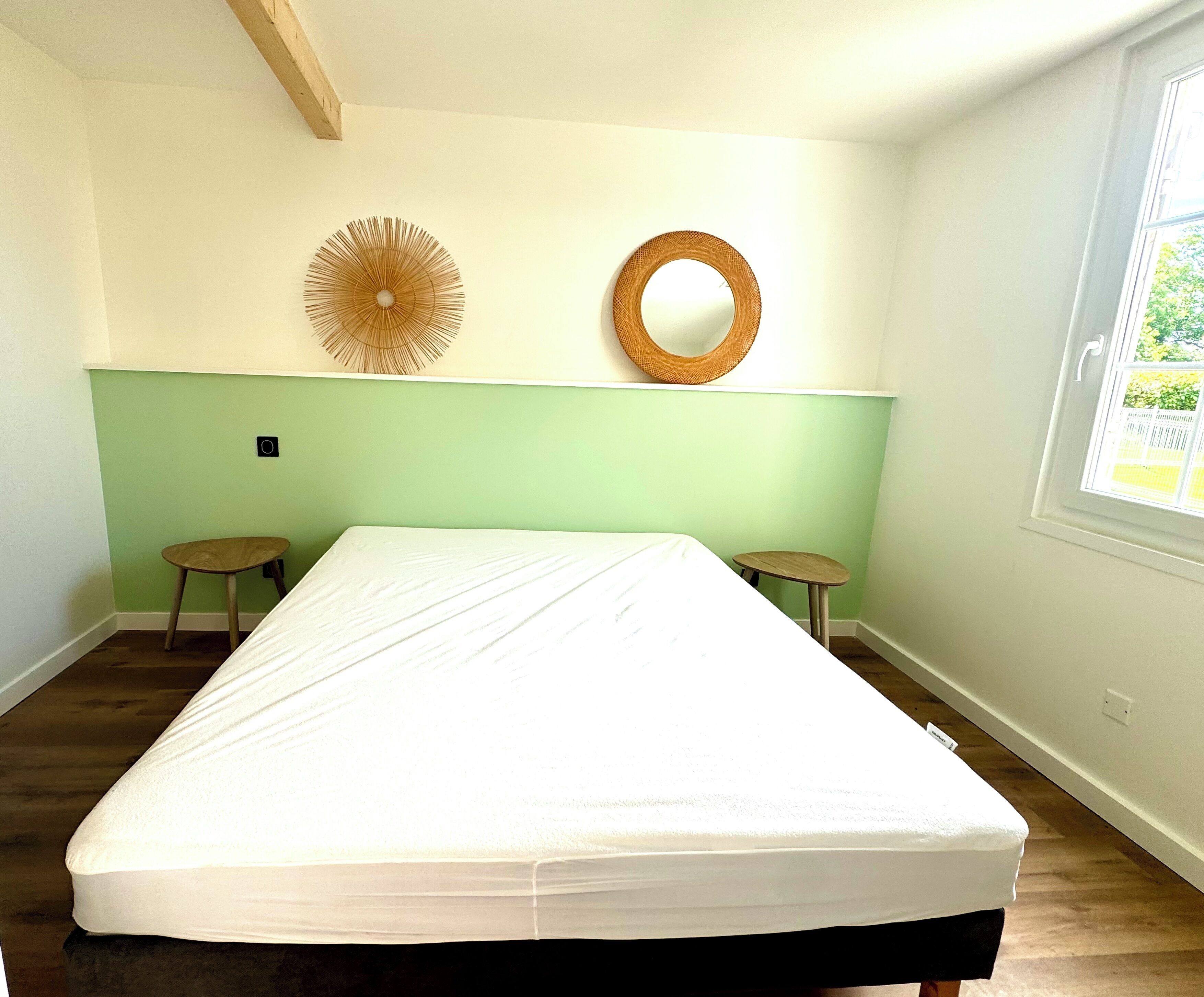 Un dormitorio con cama, dos taburetes pequeños, un espejo fijado a la pared y una ventana con vista a la vegetación.