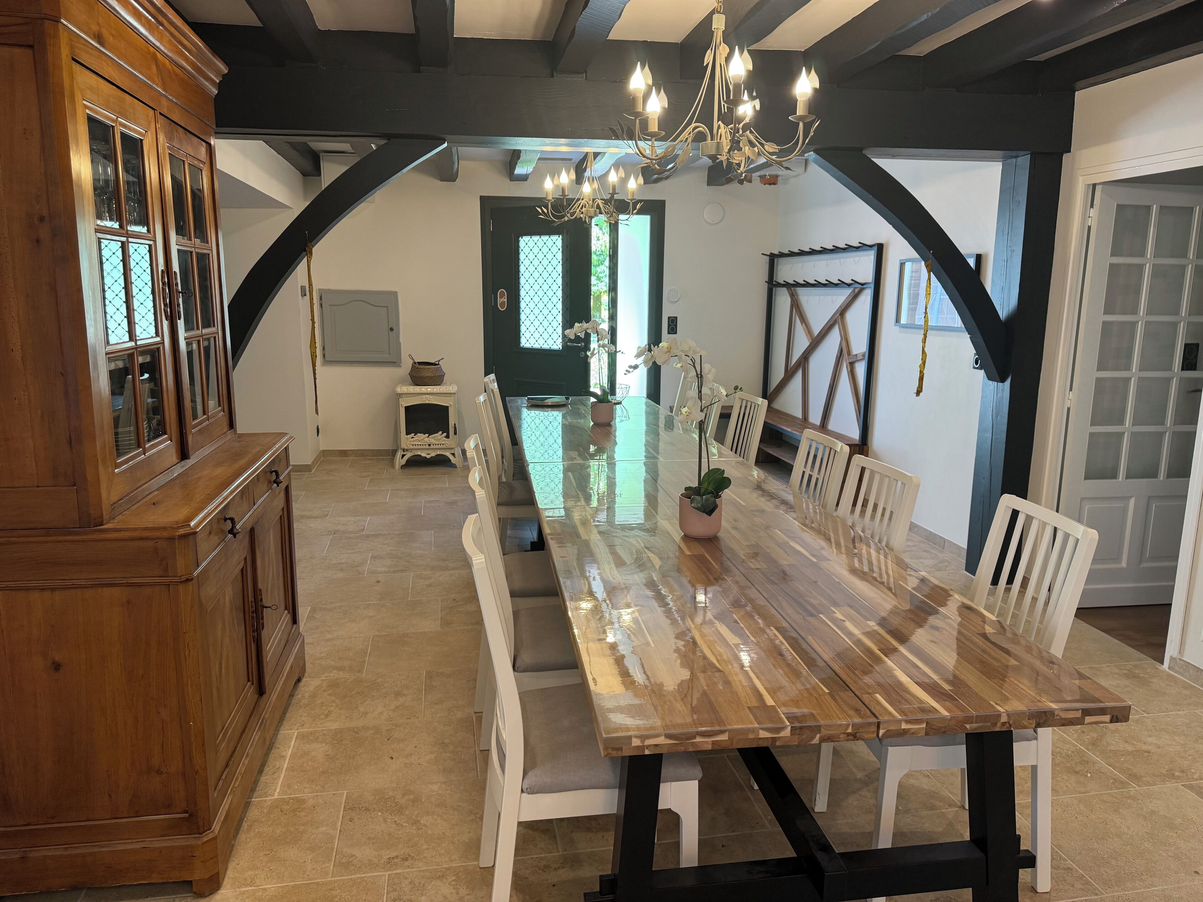 Een eetruimte met een lange houten tafel, witte stoelen, een kroonluchter en een houten kast.