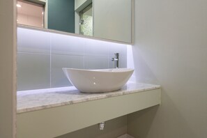 Un baño con un lavamanos blanco tipo recipiente, un espejo encima y una pared revestida de azulejos.