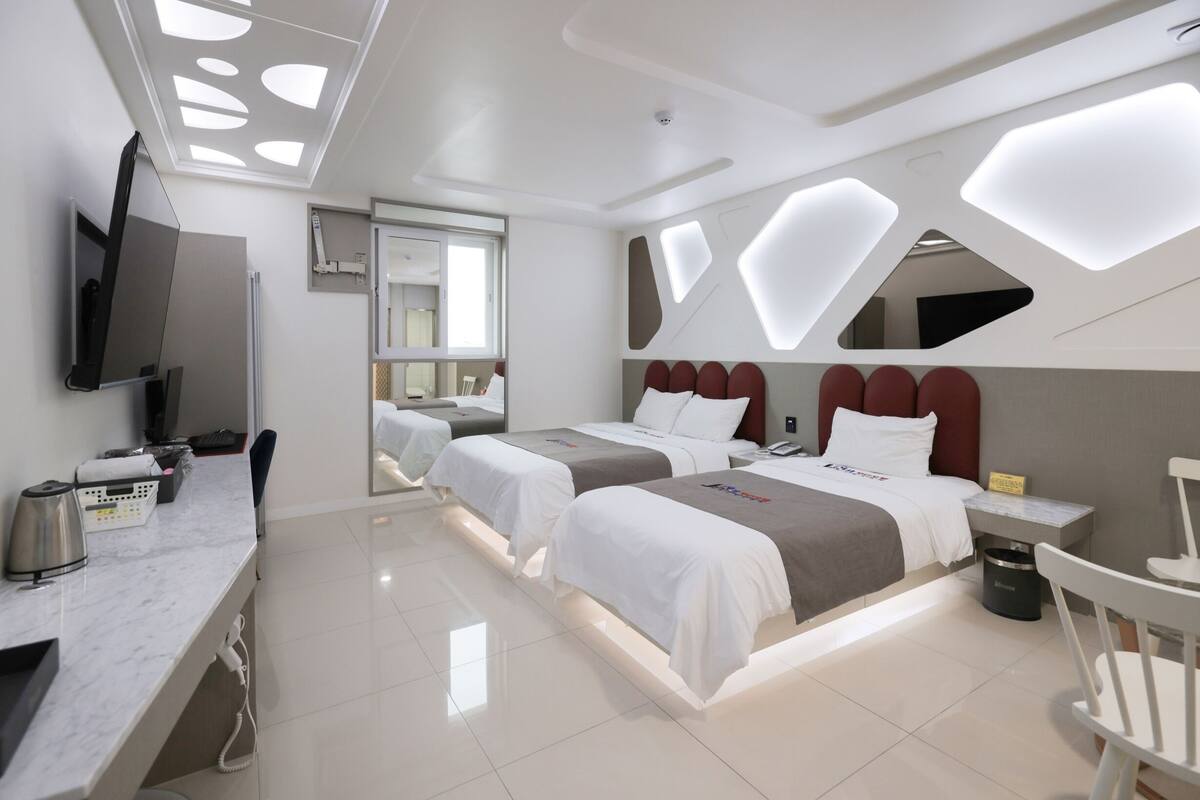 A hotel room with two beds, a desk, a chair, a TV, and a bathroom visible through a glass door.