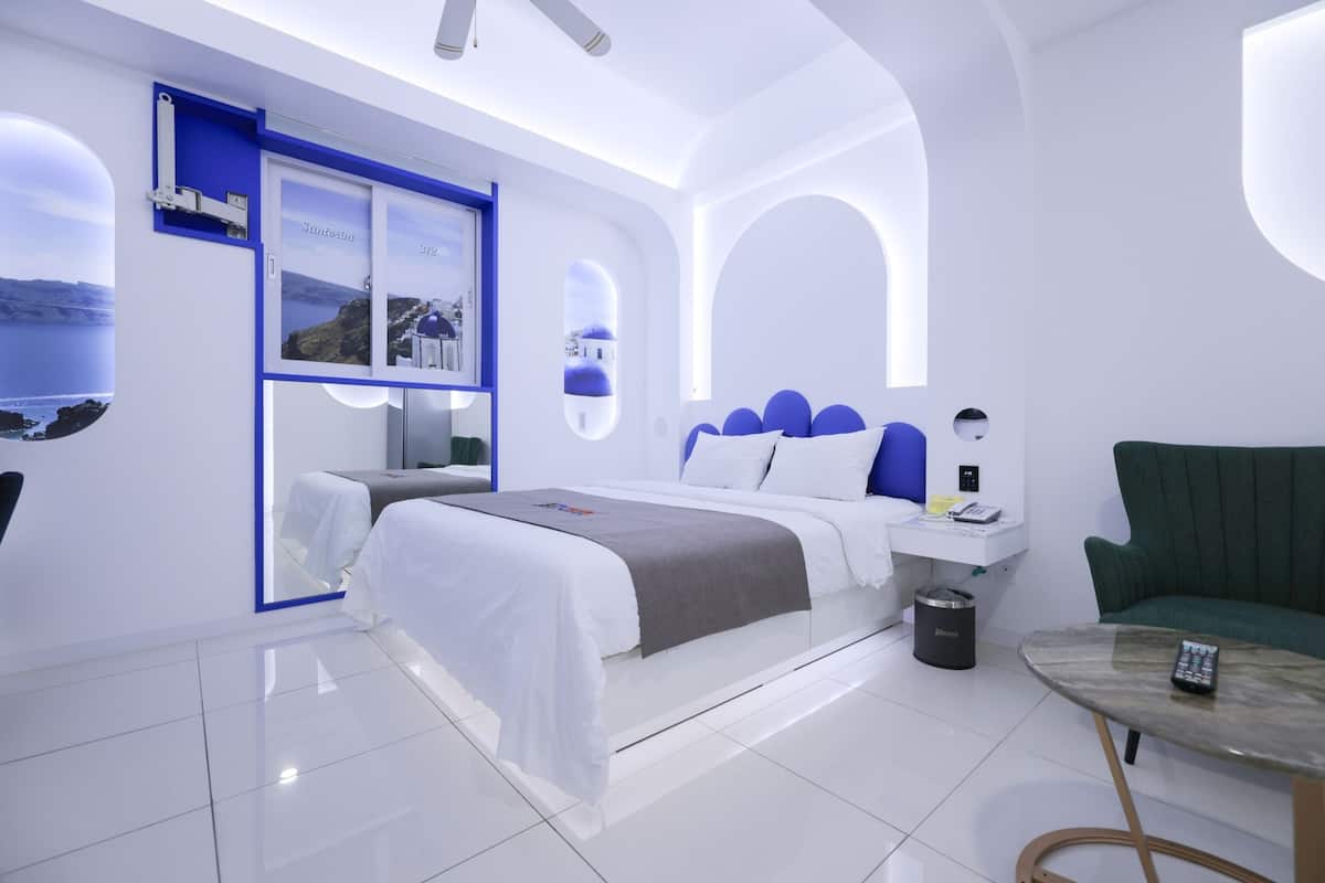 A modern hotel room with a large bed, a green armchair, a small table with a remote control, and a trash bin.
