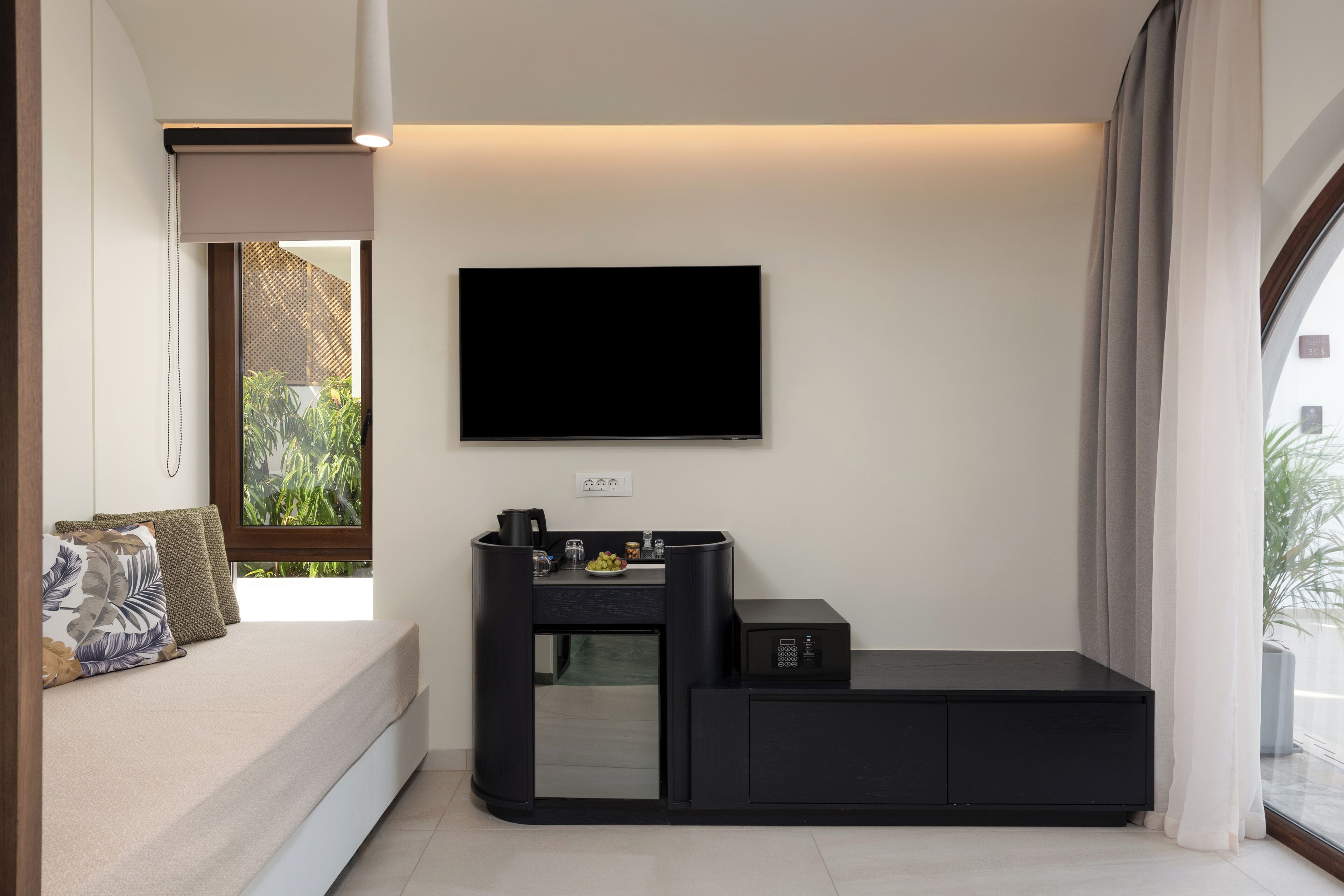 Una habitación moderna con cama, televisor de pantalla plana en la pared, pequeña zona de kitchenette y ventana con cortinas.