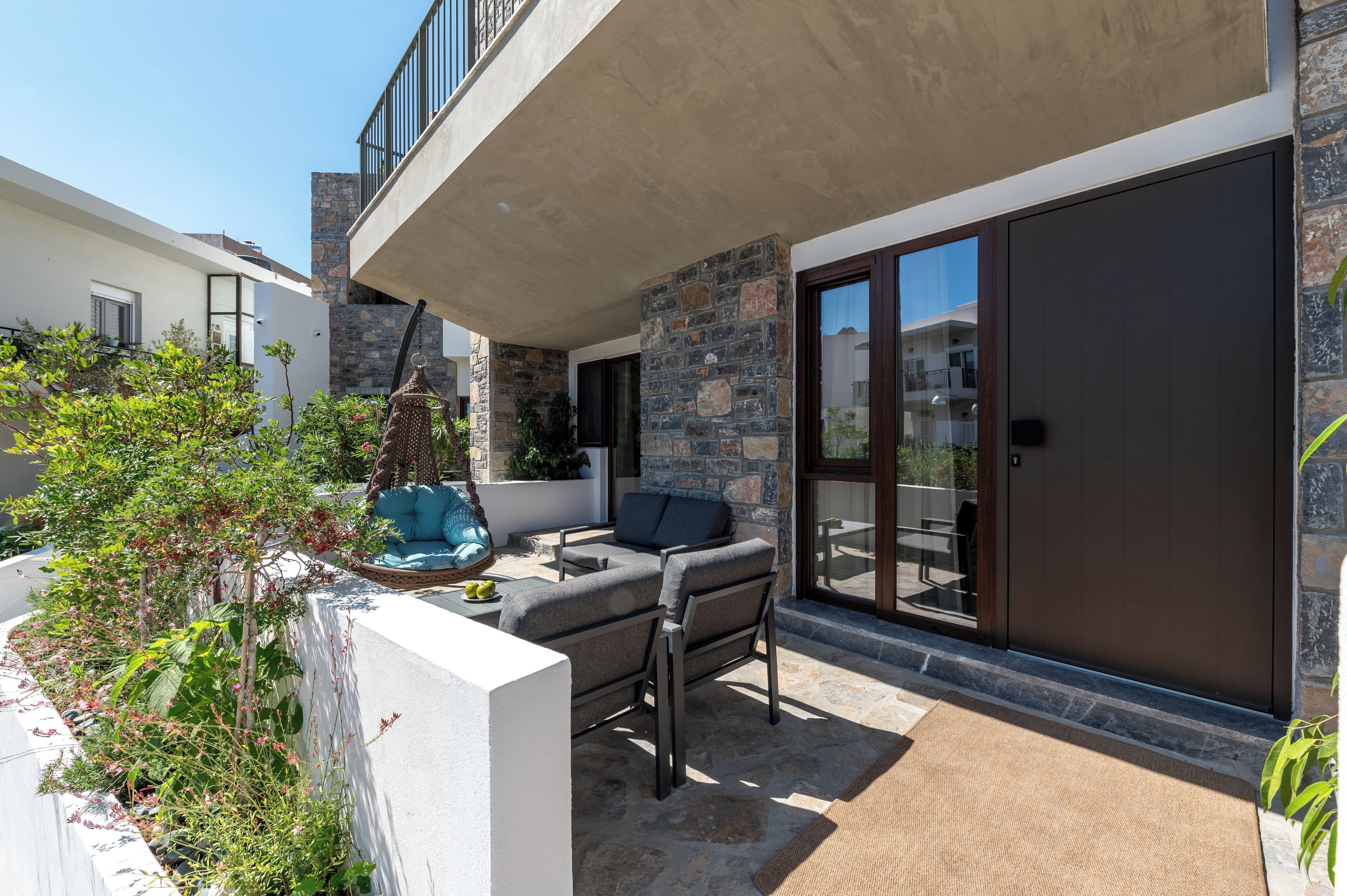Un patio moderno al aire libre con un muro de piedra, una puerta corrediza de vidrio y una zona de estar con un banco acolchado y una mesita.