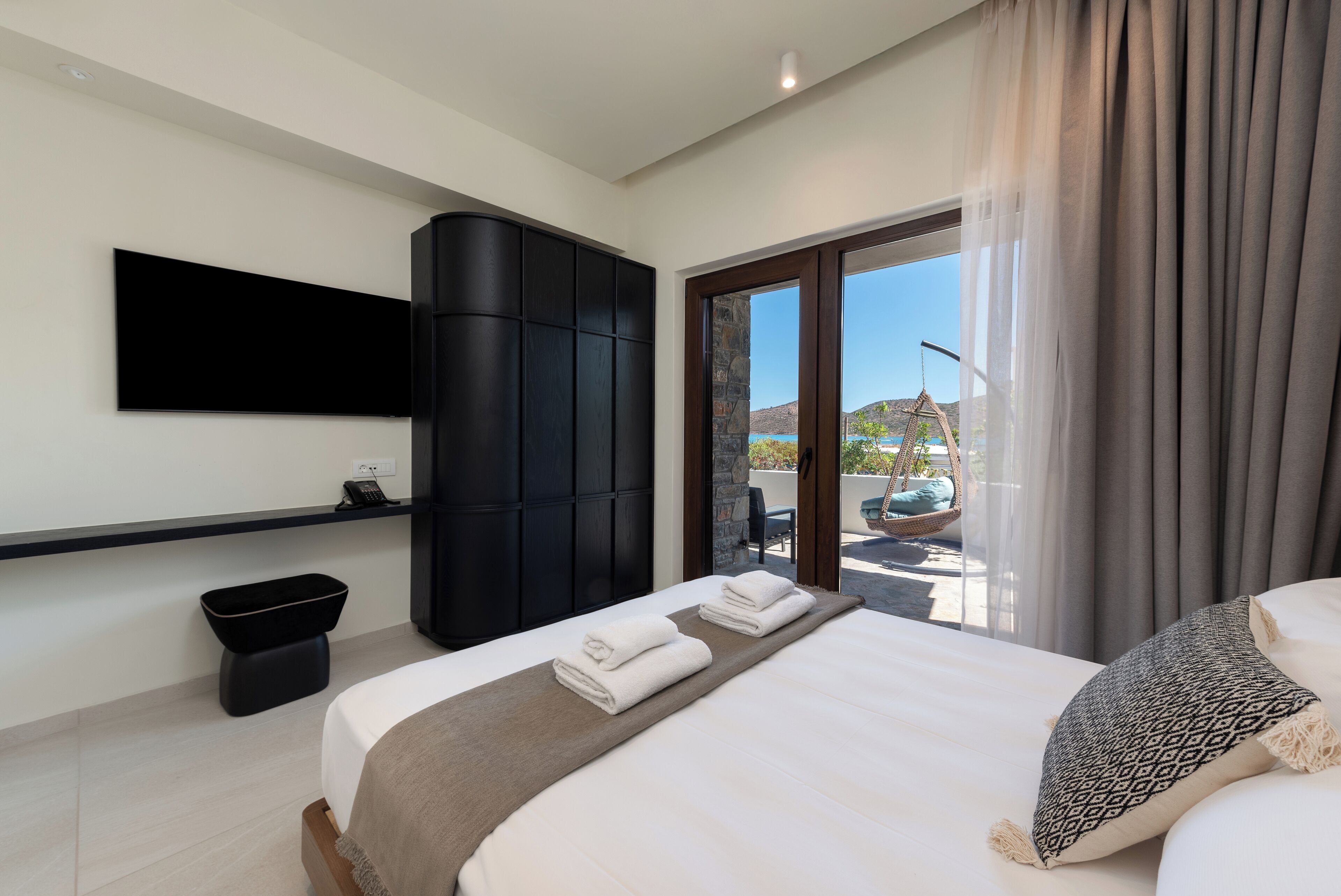 Una habitación de hotel moderna con una cama grande, un televisor de pantalla plana en la pared, un armario negro y un balcón con vistas al exterior.