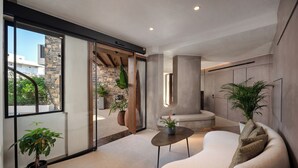 A modern interior with a large sliding glass door leading to a stone exterior, a cozy seating area with a round table, and several potted plants. - Elounda Stories Boutique Hotel (Agios Nikolaos)
