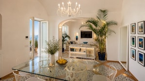 A modern dining area with a glass table, white chairs, a chandelier, and a potted plant. - Villa Mare Blu With Sea View (Praiano)