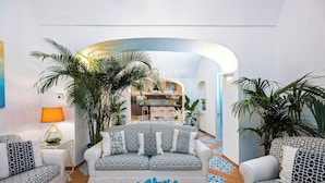 A modern living room with a sofa set, a coffee table, and a lamp. There are plants and a view of the kitchen in the background. - Villa Mare Blu With Sea View (Praiano)