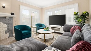 Een woonkamer met een televisie, smaragdgroene fauteuils, een salontafel en een grijze bank.