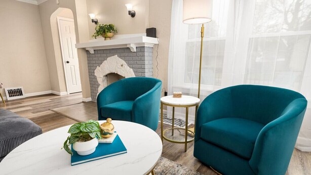 Een woonkamer met smaragdgroene fauteuils, een witte marmeren salontafel, een open haard en een groot raam met dunne gordijnen.