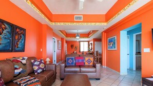 A spacious living room with a brown leather sofa, a matching ottoman, and colorful throw pillows. A large painting is visible on the left wall, and a door leads to another room.