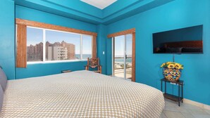 A room with a large bed, a TV mounted on the wall, a chair, and a balcony with a view of buildings.