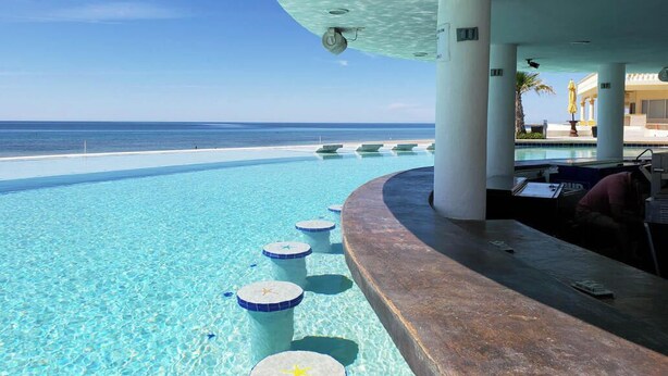 A clear blue pool with star-shaped steps, a sandy beach, and a view of the ocean.
