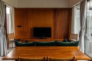 A modern living room with a large wooden TV stand, two wooden chairs, a wooden dining table, and green cushions.